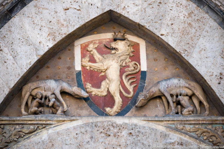 Pointed stone tympanum with a crowned lion on a red shield, flanked by two reclining carved animals.