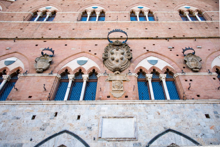 Brick Palazzo Pubblico façade with pointed Gothic windows, a large central coat of arms, smaller heraldic shields, and stone plaques.