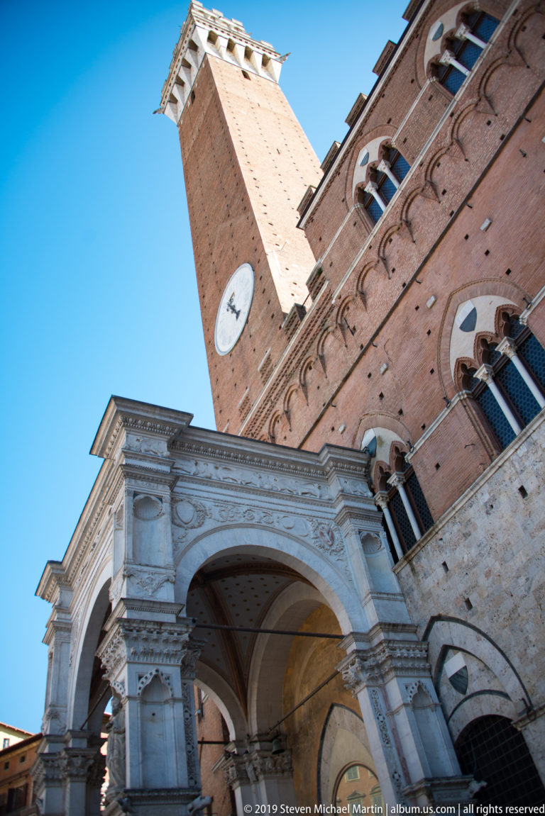 Low-angle view combining Cappella di Piazza, Palazzo Pubblico’s brick façade, and the soaring Torre del Mangia against blue sky.