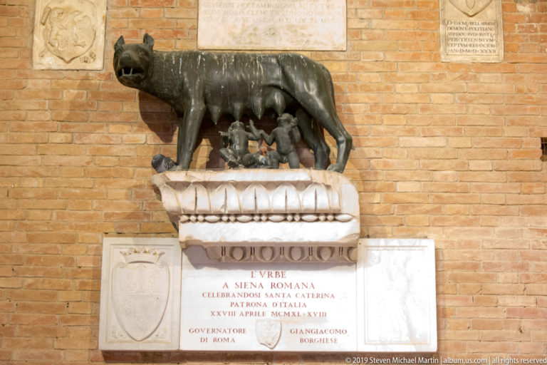 Bronze she-wolf nursing Romulus and Remus on a stone pedestal above inscribed marble plaques mounted on a brick wall.