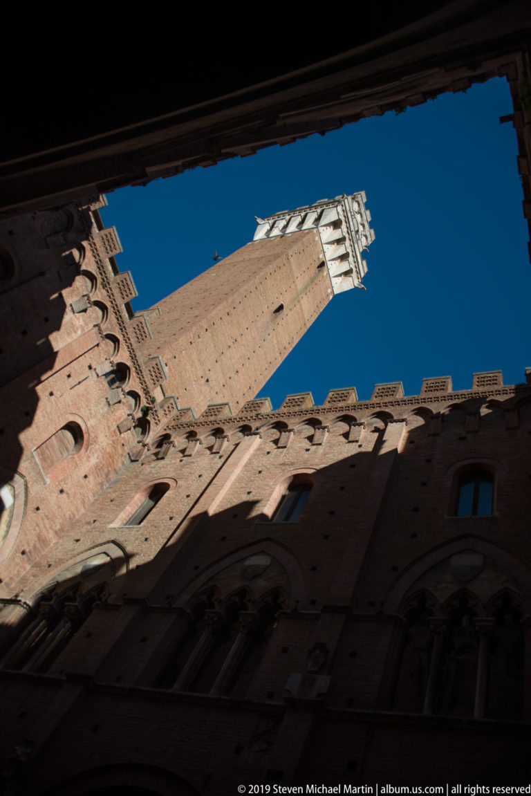 Steep upward view of Torre del Mangia and its pale belfry framed by shadowed crenellated brick walls against blue sky.