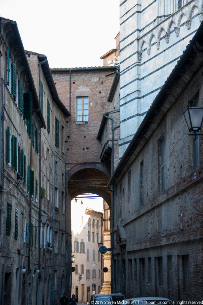 Narrow Siena street with weathered masonry, green shutters, and an enclosed brick bridge spanning overhead toward sunlit buildings beyond.