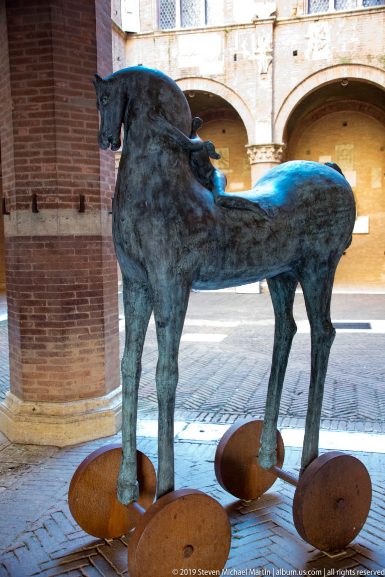 Dark stylized metal horse standing on four rust-colored circular supports beneath brick arcades in Siena.