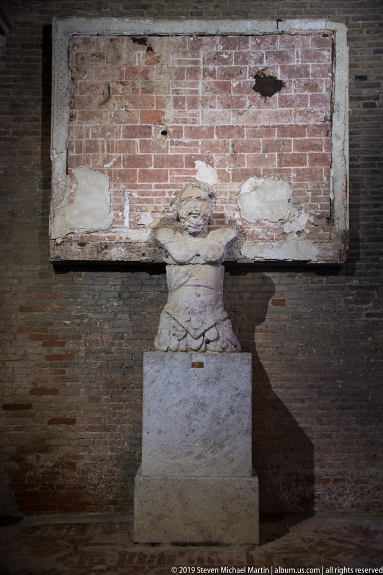 Headless fragmentary stone figure on a pedestal against a brick wall with a large rectangular patch of exposed masonry.