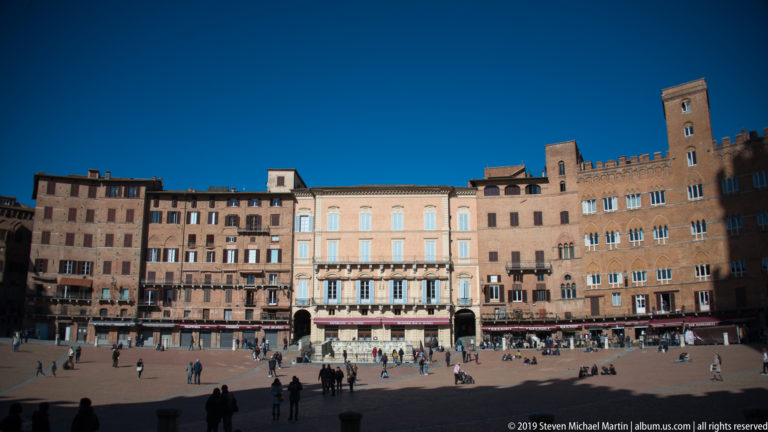 Sunlit row of ochre and brick palazzi lining Piazza del Campo, with visitors scattered across the sloping brick square.