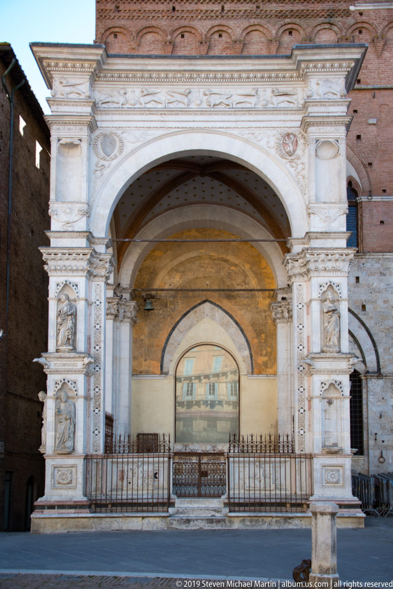 Open marble Cappella di Piazza with broad arches, sculpted niches, carved piers, and brick Palazzo Pubblico behind.
