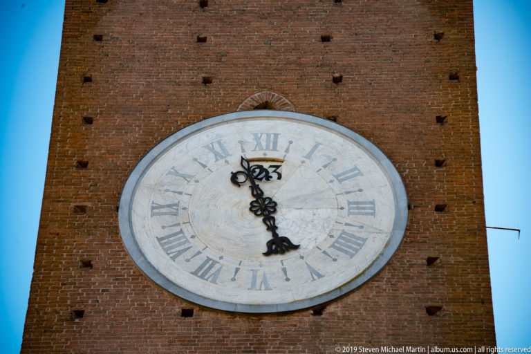 Large pale circular clock with Roman numerals and ornate black hands mounted on the red-brick Torre del Mangia.
