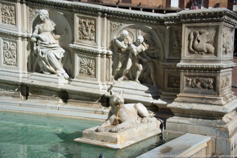 Reclining stone she-wolf on a platform in green fountain water before richly carved marble relief panels.