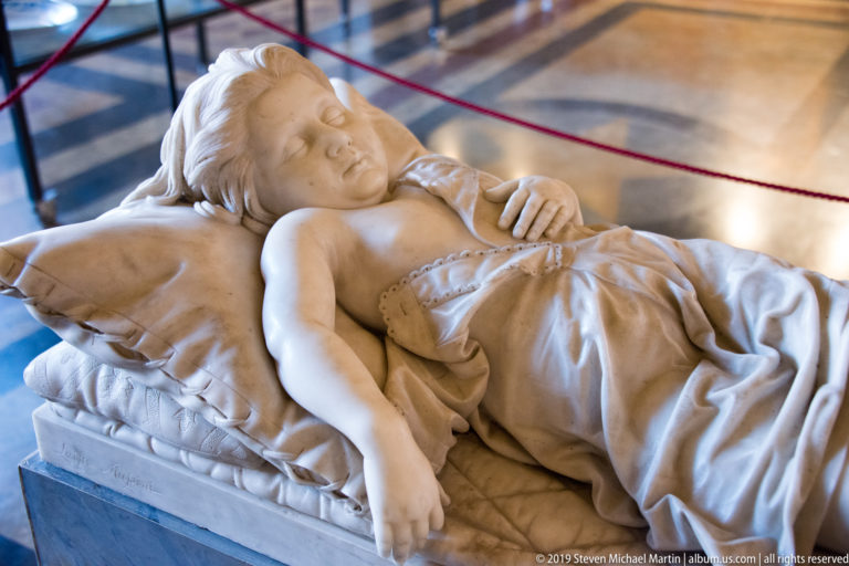 Marble sculpture of a sleeping child lying on pillows and rumpled bedding, with one arm hanging over the mattress edge.