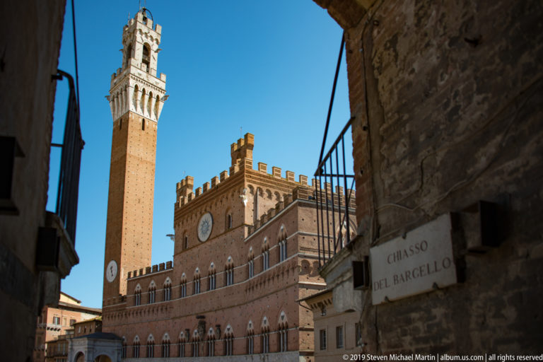 Sunlit Palazzo Pubblico and Torre del Mangia framed by the dark Chiasso del Bargello passage and its wall sign.