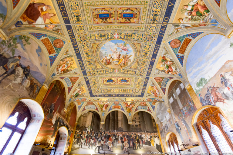 Wide decorated hall with elaborate painted ceiling, arched inscriptions, historical crowd murals, and tall windows along both sides.