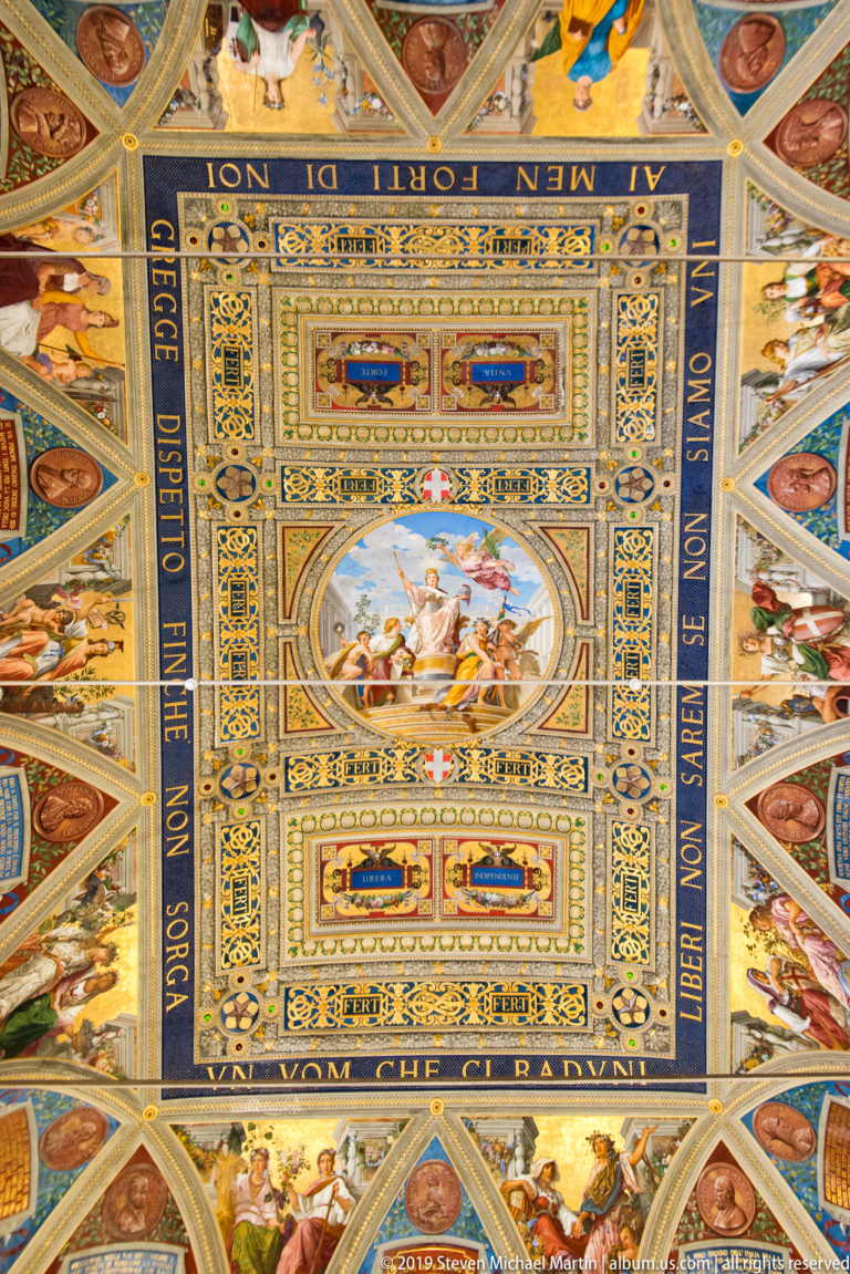 Centered view of an ornate ceiling with oval figural painting, blue inscription bands, gilded ornament, portraits, and surrounding figures.