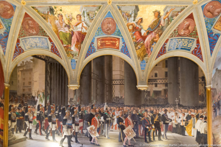 Large historical mural of uniformed participants, clergy, banners, and spectators beneath three decorated arches with inscriptions above.