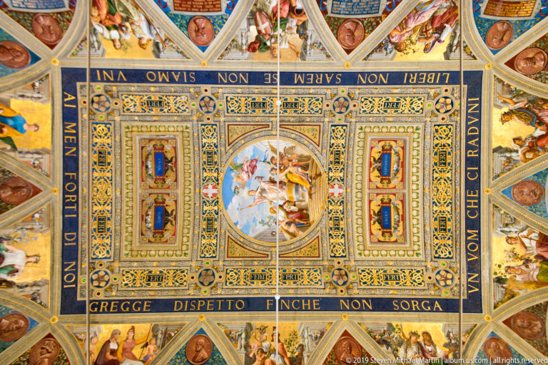 Ornate rectangular ceiling with Italian inscriptions, gilded borders, portrait medallions, and a central oval allegorical painting.