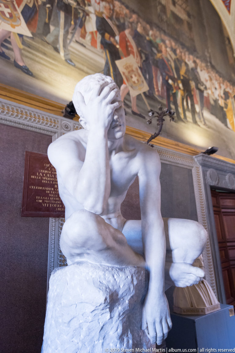 White marble male figure reclining on a rough support, head resting on one hand, with a historical mural behind.
