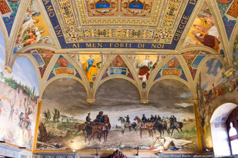 Large wall mural of mounted troops crossing a landscape beneath painted arches, inscriptions, and ornate ceiling decoration.