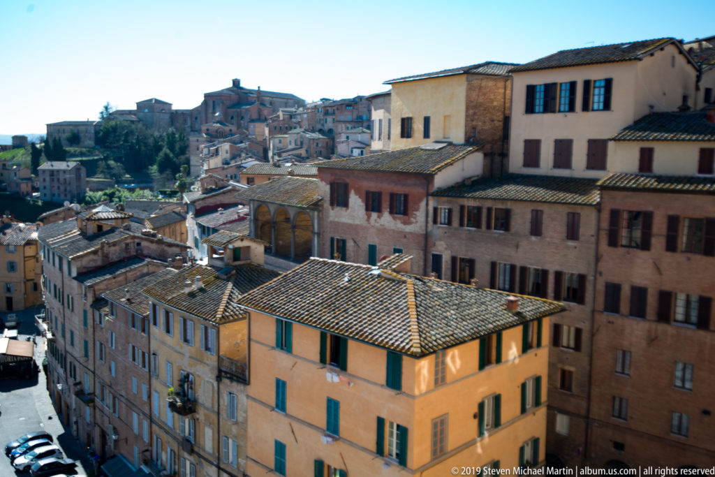 Elevated Siena neighborhood with terracotta roofs, ochre and brick façades, shuttered windows, and buildings climbing the hillside.
