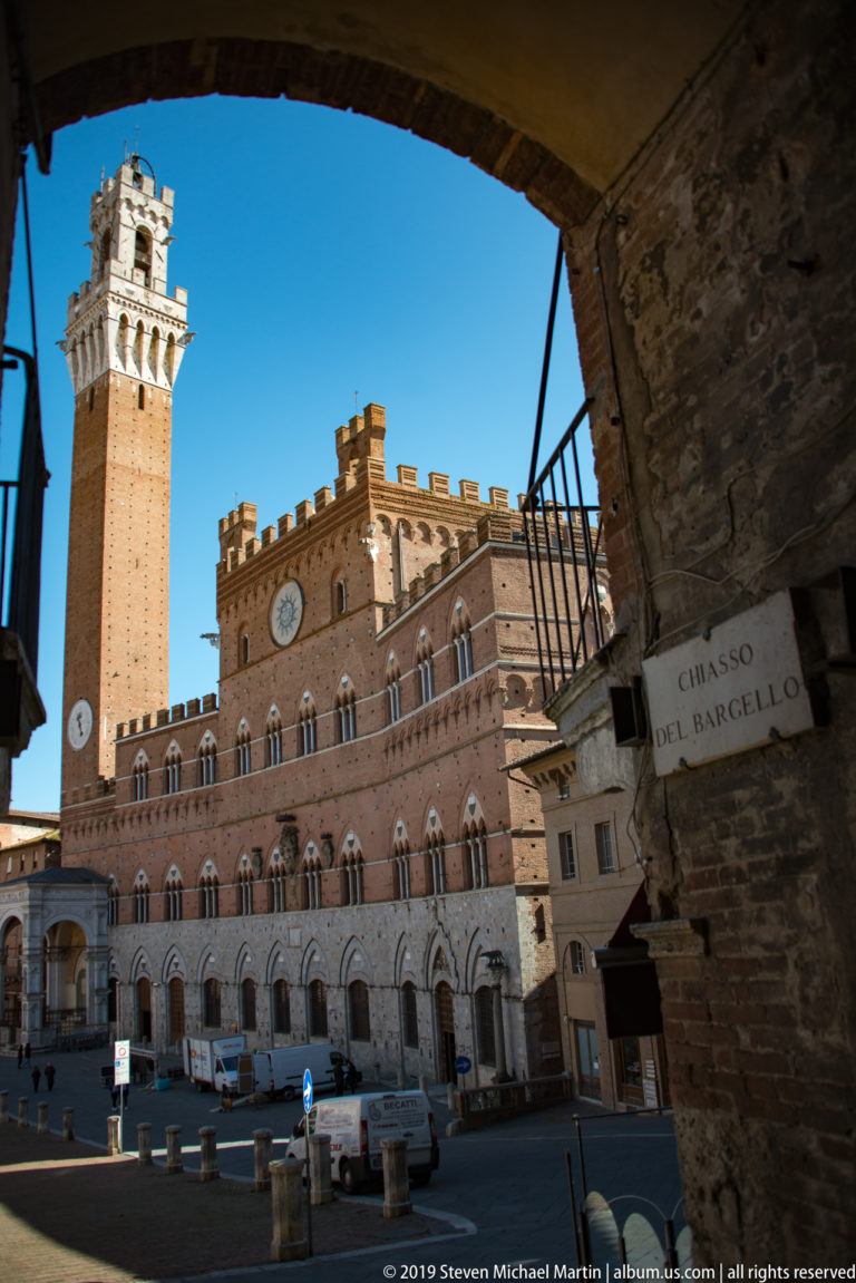 Palazzo Pubblico and Torre del Mangia seen through the shaded Chiasso del Bargello archway, with the passage sign at right.