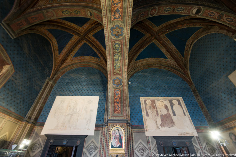 Blue star-patterned rib vault above fragmentary wall frescoes, with painted ribs and a small devotional panel between larger damaged areas.