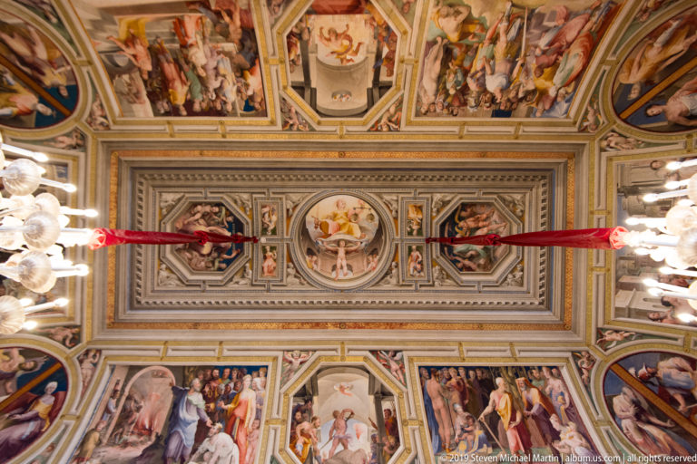 Ornate painted ceiling centered on a circular medallion, surrounded by framed narrative scenes, decorative figures, and geometric borders.