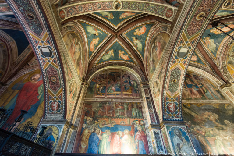 Upward view through a rib-vaulted chapel densely painted with saints, angels, narrative scenes, and ornamental borders.