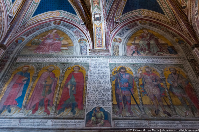 Painted wall with standing haloed saints flanking a tall inscribed panel beneath two arched narrative scenes.