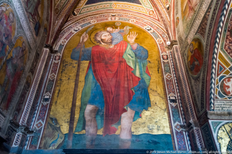 Monumental fresco of Saint Christopher in red and blue carrying the Christ Child on his shoulder and holding a tall staff.