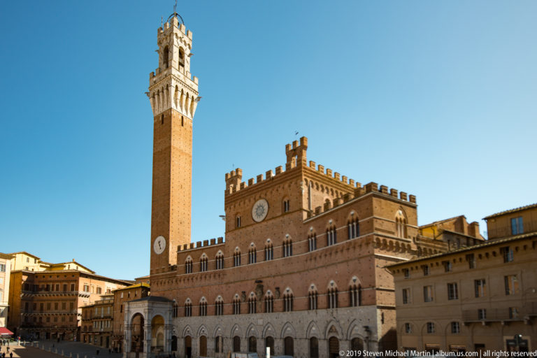 Close exterior view of Palazzo Pubblico with pointed windows, crenellations, pale arcade, clocks, and Torre del Mangia rising above.
