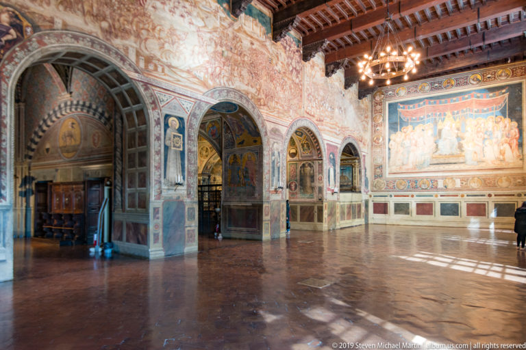 Oblique interior view with timber ceiling, tiled floor, decorated arches, faded wall frescoes, and a large sacred mural at the far end.