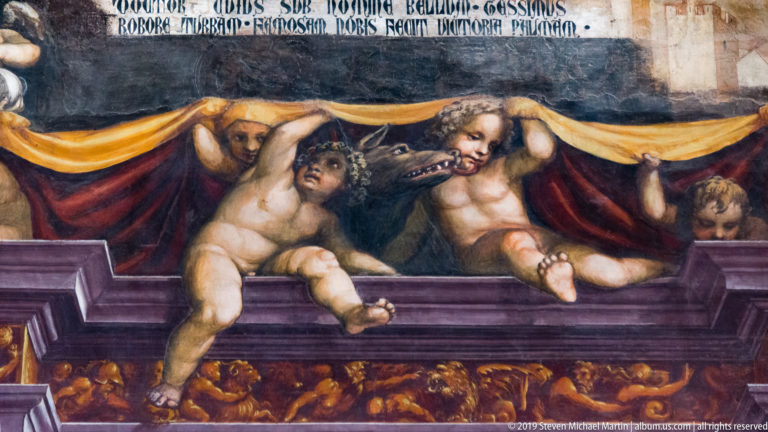 Two nude winged figures pull back a red-and-gold curtain above a dark lower scene crowded with smaller figures.