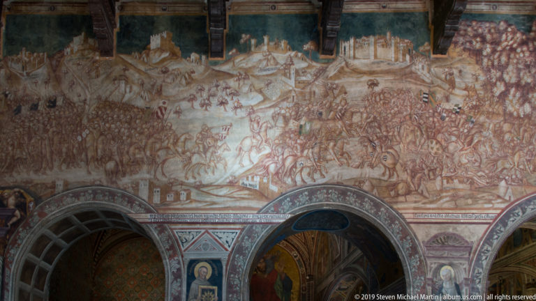 Large faded battle fresco filled with mounted troops and infantry above a lower register of decorated arches and painted panels.