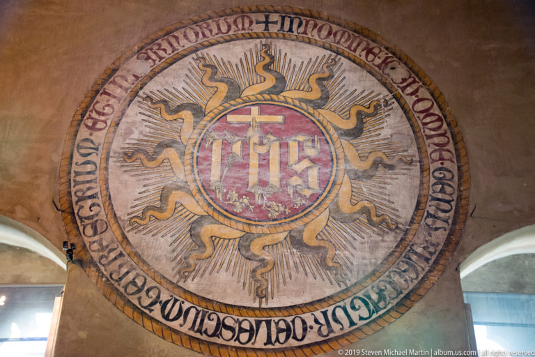 Large circular IHS Christogram with red center, radiating pale rays, curling golden flames, and a painted inscription around the border.