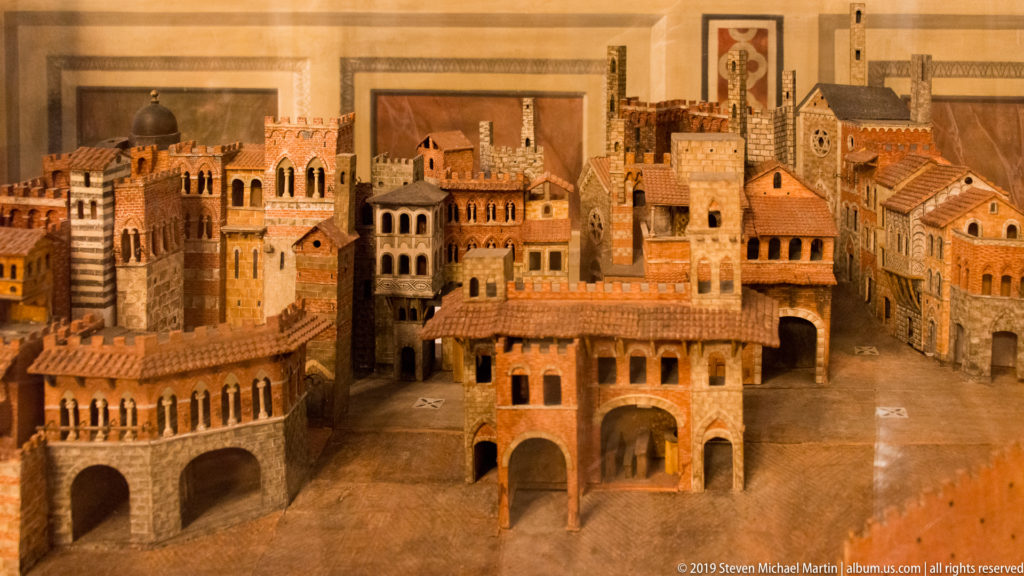 Detailed architectural model of a medieval city with towers, arcades, narrow streets, striped masonry, and terracotta roofs.