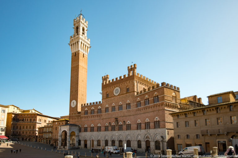 Palazzo Pubblico’s crenellated brick façade and arcaded base beneath the tall Torre del Mangia against a clear blue sky.