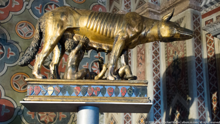 Gilded she-wolf standing over two nursing infants on a patterned support against a richly decorated wall.
