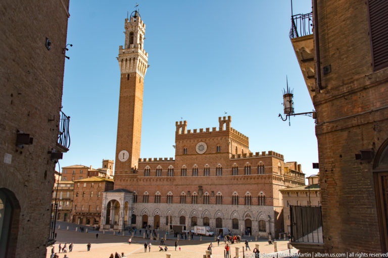 Palazzo Pubblico and tall Torre del Mangia in sunlight across Piazza del Campo, framed by shadowed buildings on both sides.