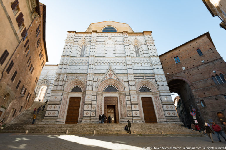 Frontal view of a large striped marble façade with three pointed portals, geometric panels, and a plain upper wall.