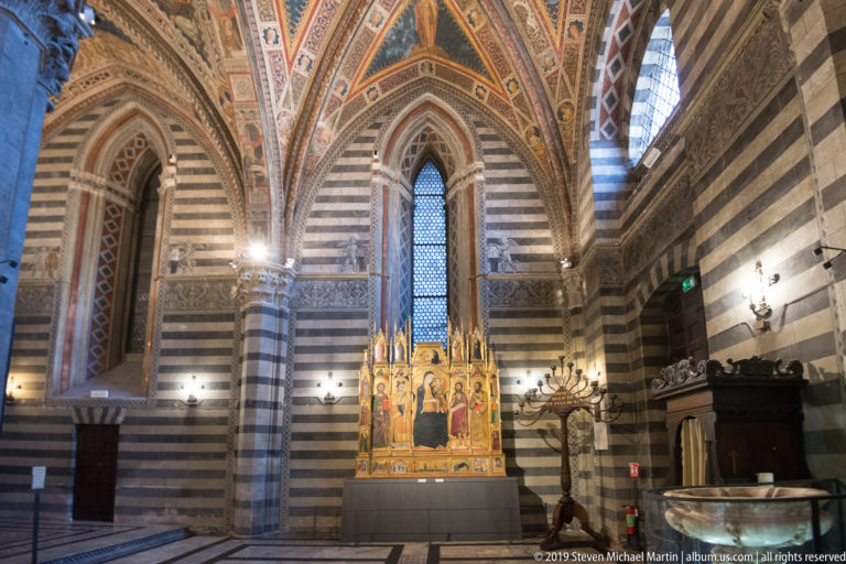 Striped Gothic interior with tall pointed arches, narrow windows, and a gilded multi-panel altarpiece centered below.