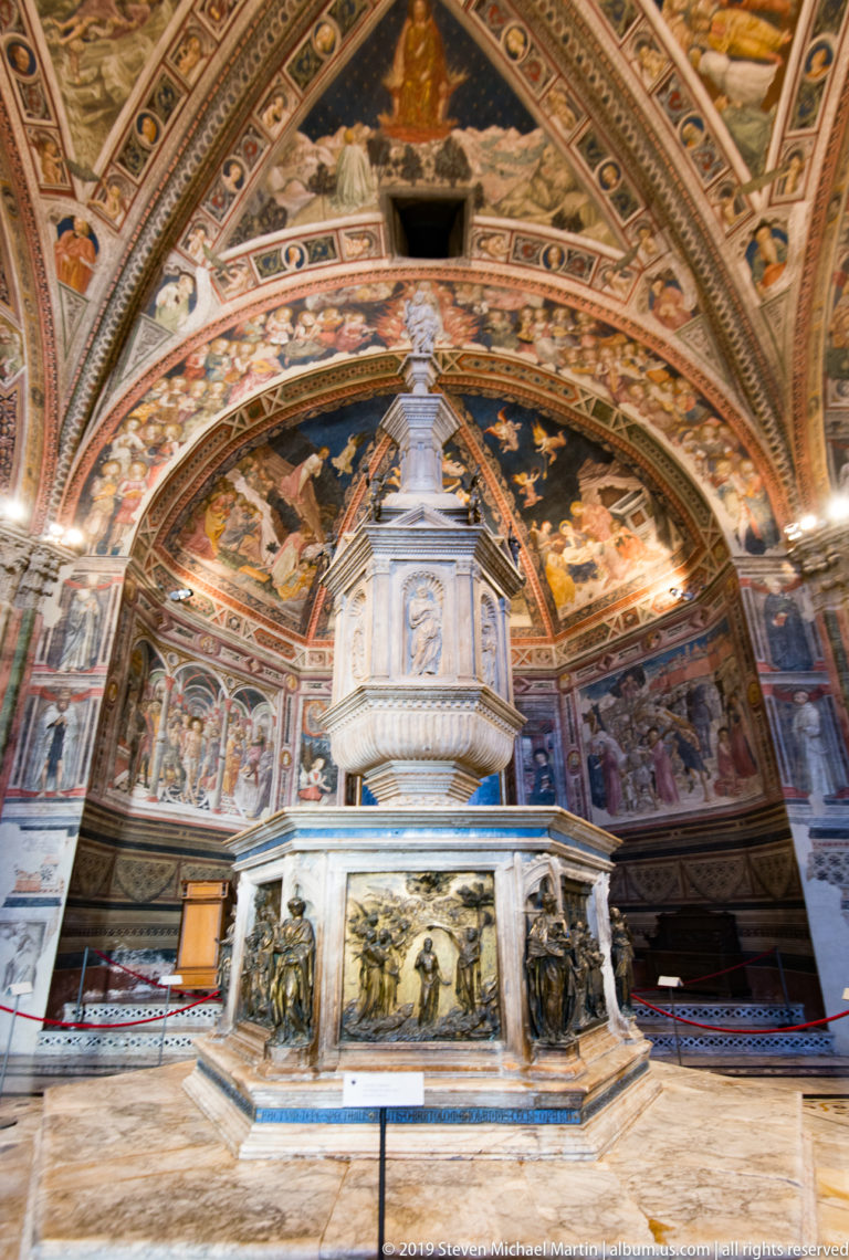 Frontal view of a pale marble monument with dark relief panels on its polygonal base, set before a fresco-covered apse.