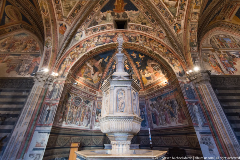 Wide frontal view of a tall white marble monument centered before a frescoed apse beneath a richly painted vault.