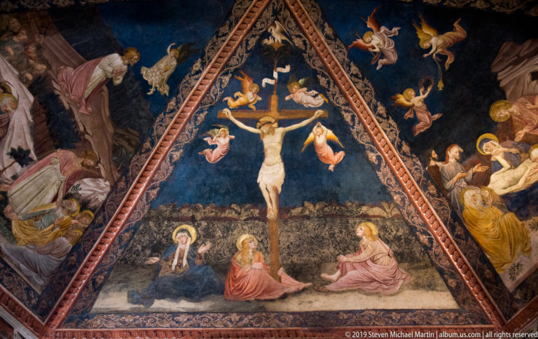 Crucifixion fresco with Christ on the cross, angels above, mourners below, and painted figures filling a triangular vaulted compartment.