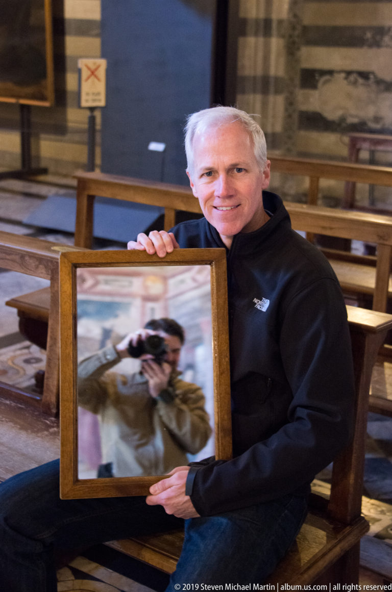 Seated man smiling while holding a framed reflective image of the photographer inside a striped-masonry church interior.