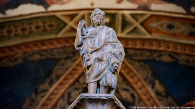 Small bearded marble figure standing above a shell-shaped basin, one hand raised, against richly painted chapel frescoes.