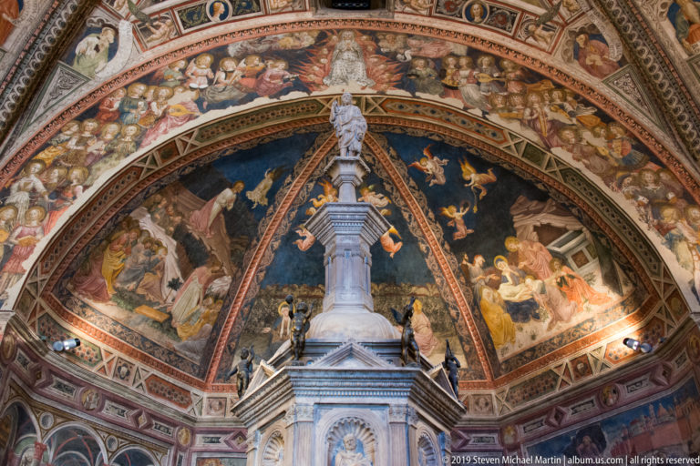 Fresco-covered apse with a tall central white marble monument, segmented painted vault, and narrative scenes across the curved walls.