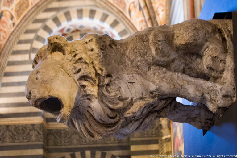 Close view of a weathered carved stone animal head with open mouth and curling mane, projecting from decorated architecture.