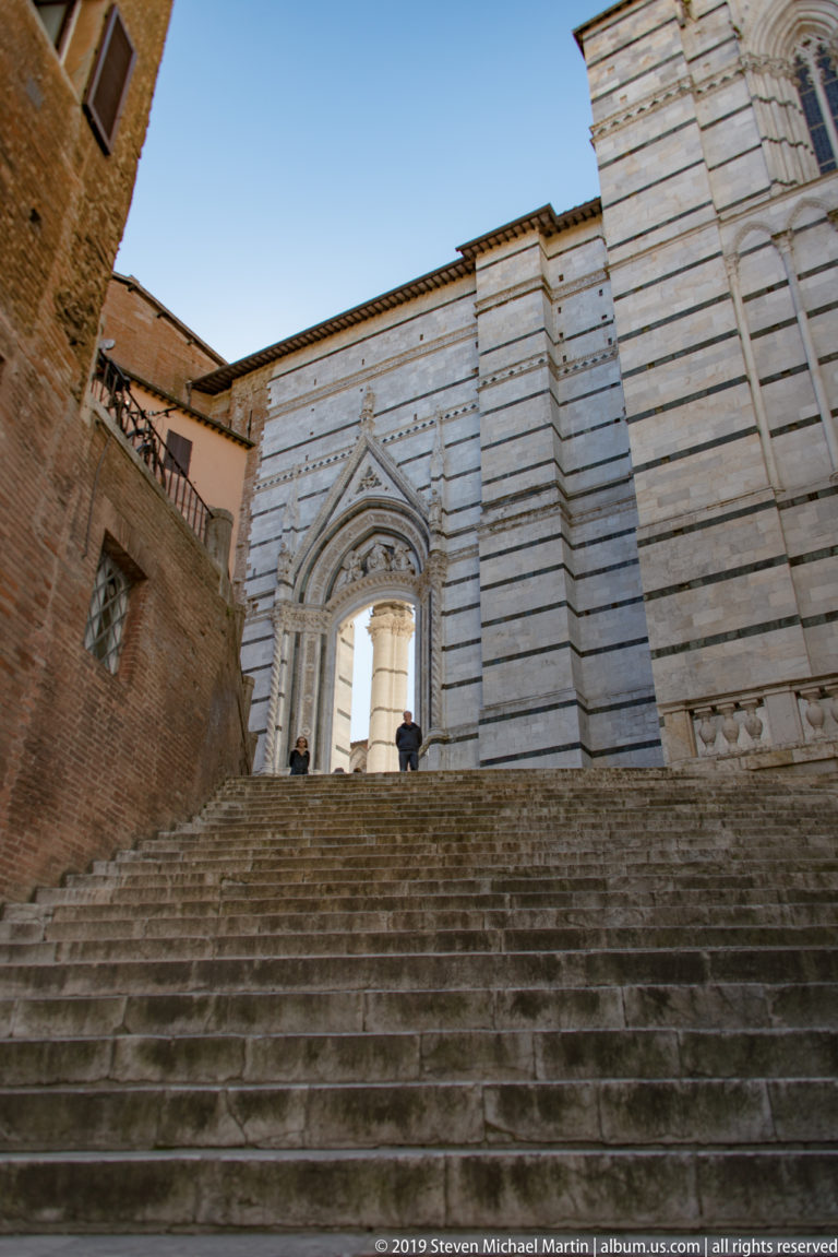 Wide stone stair rising to a striped marble façade with three pointed Gothic portals and geometric lower-wall decoration.
