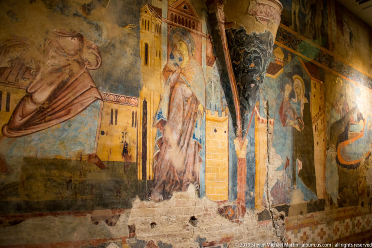 Fragmentary medieval frescoes with saints, buildings, patterned borders, and narrative scenes wrapping around an interior corner.