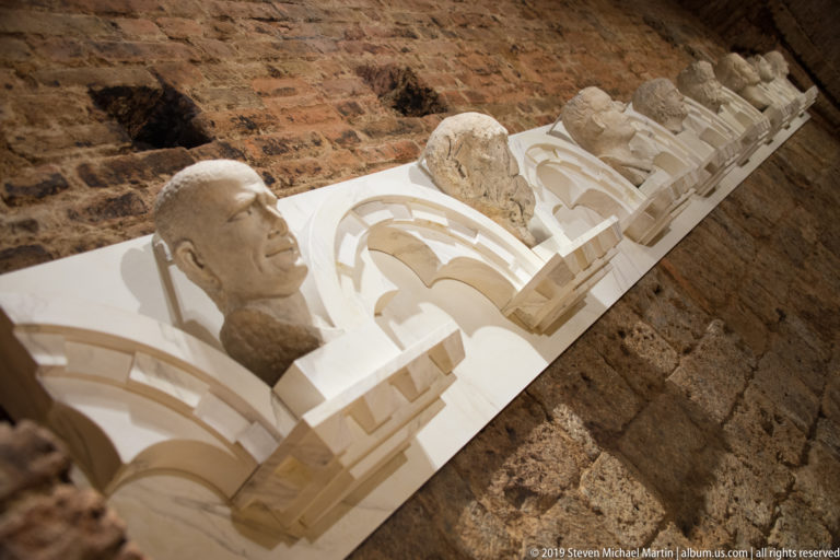 Diagonal display of carved stone heads set among reconstructed white Gothic tracery against an exposed brick wall.