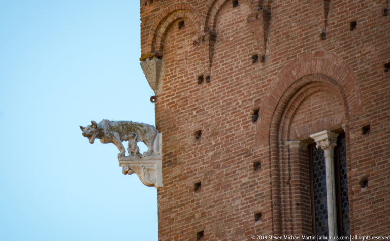 Projecting she-wolf and twins on a carved stone bracket at the corner of Palazzo Pubblico, silhouetted against blue sky.