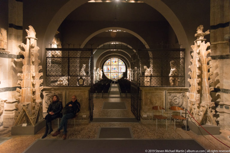 Two seated visitors beside carved stone fragments, with a metal screen and arched sculpture gallery leading to circular stained glass.